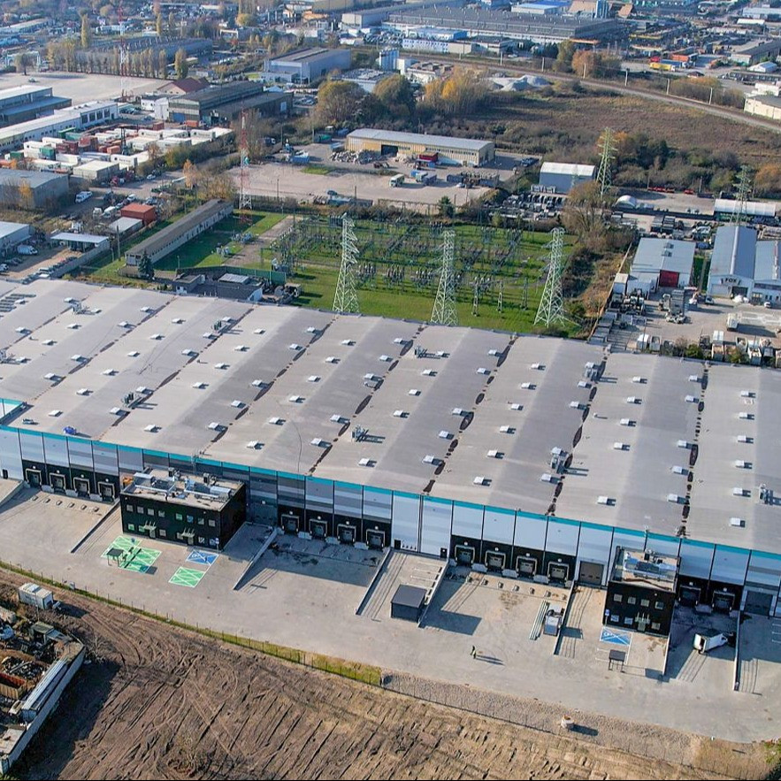 Nowoczesny budynek Gdynia City Logistics z dużą halą magazynową i częścią biurową, widoczny z zewnątrz. Na pierwszym planie plac manewrowy i doki załadunkowe.